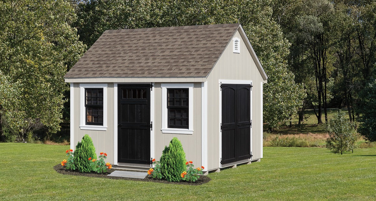 Small gray storage shed with black doors and windows on a grassy lawn with trees in the background.