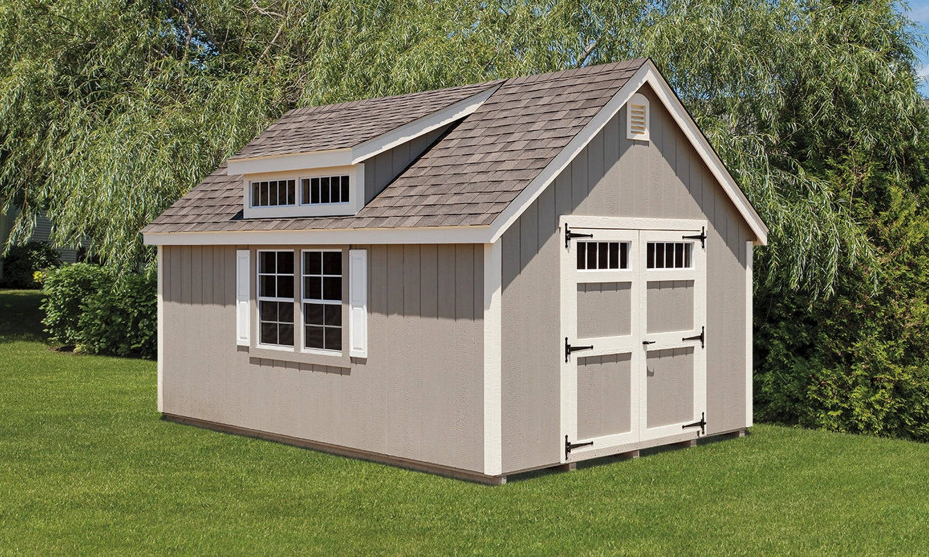 Small storage shed with a gray roof and white trim on a grassy area with trees in the background.