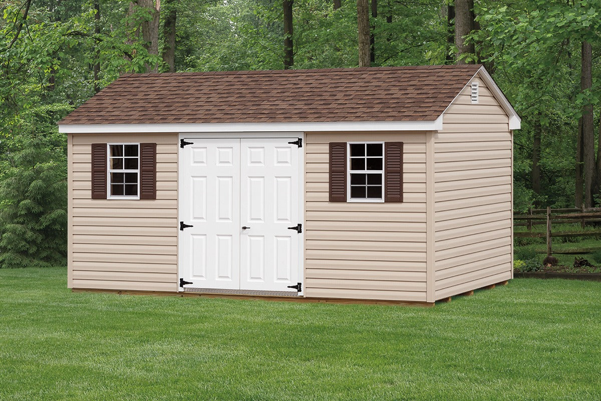 Wooden storage shed with a brown roof and white doors on a grassy area with trees in the background.